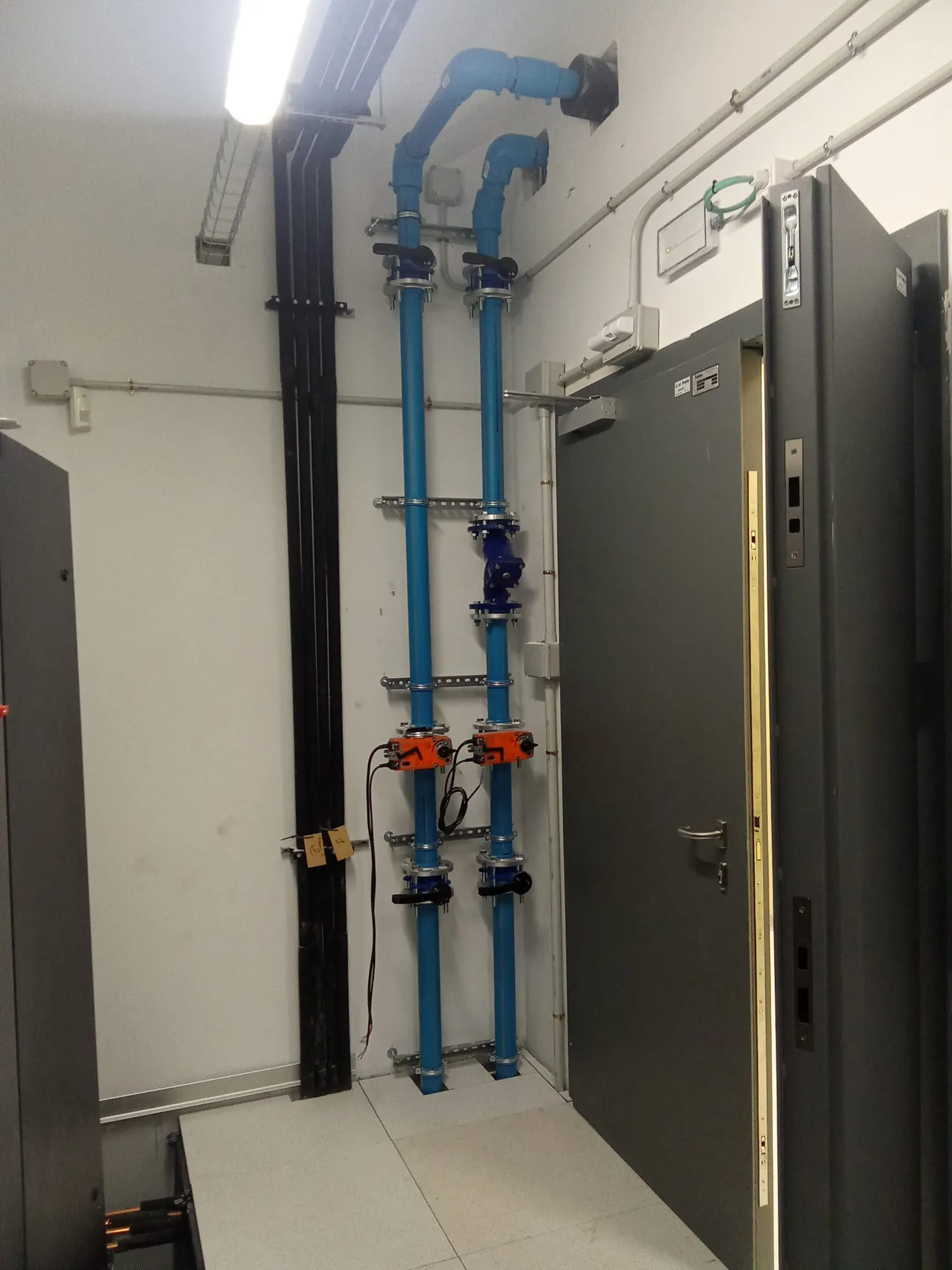 Montante de tuberías azules con válvulas de zona junto a la puerta de una sala técnica
